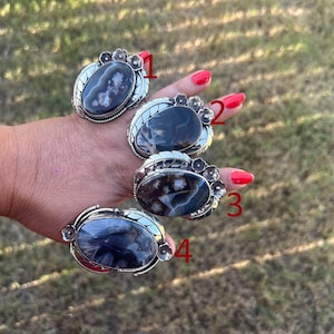 May include: Four silver rings featuring large oval-shaped dark gemstones with unique white and grey inclusions. Each ring is accented with small silver flower motifs on the sides and numbered one through four in red text for identification.