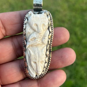 Könnte beinhalten: Elfenbeinfarbener Anhänger mit einem geschnitzten Design eines Bären und eines Wolfes. Der Anhänger ist in einem silberfarbenen Metallrahmen mit dekorativem Rand eingefasst. Der Anhänger ist rechteckig und wird von einer Hand gehalten.