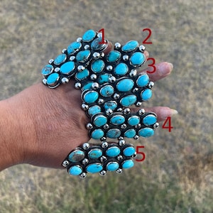 May include: A silver cuff bracelet featuring five rows of oval-shaped turquoise stones set in metal bezels. The stones vary in shades of blue and green with dark matrix patterns. The numbers 1 through 5 are printed in red next to each row.