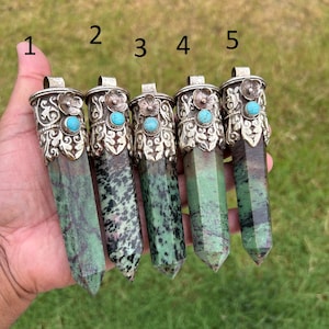 Op de afbeelding: Vijf groene en zwarte edelsteenhangers met zilverkleurige metalen doppen. Elke dop heeft een bloem en een turkooizen steen. De hangers worden in een hand gehouden tegen een vage groene achtergrond.