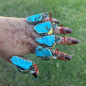 May include: Five silver rings with large, irregular turquoise stones are displayed. Each ring has a unique design, with the turquoise stones varying in shape and color. The rings are set against a green background.