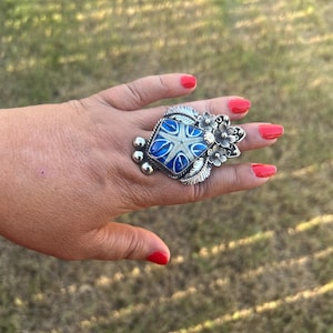 May include: A silver ring featuring a central square inlay with a blue and white starfish design. The setting is decorated with metal floral accents, leaves, and small silver spheres along the side, creating a detailed metallic appearance.