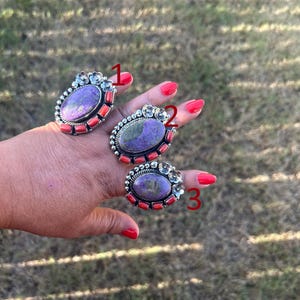 May include: Three silver rings labeled 1, 2, and 3, each featuring an oval purple stone set in a metal frame. The rings are accented with small orange coral-like pieces and decorative silver floral elements along the top edges of the settings.