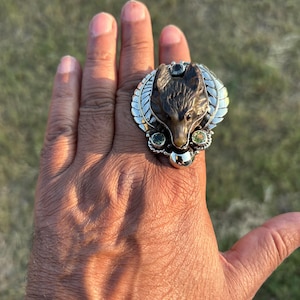 May include: A silver ring featuring a detailed wolf head with a dark brown finish. The ring has a decorative design with green and silver accents. The ring is worn on a hand, showcasing its size and design.