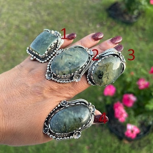 May include: Four silver rings featuring large, oval and rectangular green rutilated quartz stones with dark needle-like inclusions. Each ring is set in a decorative silver bezel with small metal bead and floral accents, numbered one through four.