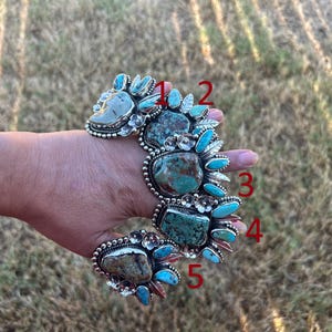 May include: A wide silver cuff bracelet featuring five large, irregular turquoise stones set in ornate metalwork. The design includes smaller turquoise accents, silver leaf motifs, and small flower details. Red numbers 1 through 5 are visible.