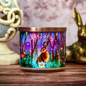May include: A decorative candle with a copper-colored lid and a colorful forest scene featuring a rabbit, the word "Ostara", and other forest creatures. A gold-colored rabbit statue and a decorative candle holder are also in the image.