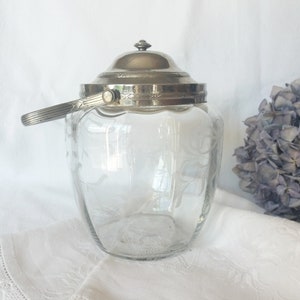 Op de afbeelding: Een glazen pot met een zilveren deksel en een zilveren handvat. De pot heeft een gegraveerd bloemenmotief in het glas. De pot staat op een wit tafelkleed.