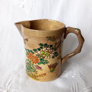 Peut inclure: Pichet en céramique vintage avec une anse en forme de bambou. Le pichet beige est décoré de motifs floraux et d'insectes colorés. Les fleurs sont dans les tons orange, blanc et vert. La poignée est marron avec des accents verts.
