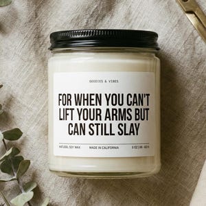 May include: A soy wax candle in a glass jar with a black lid. The label reads "FOR WHEN YOU CAN'T LIFT YOUR ARMS BUT CAN STILL SLAY." A gold-colored wick trimmer, matches, eucalyptus, and lavender sprigs are arranged around the candle.