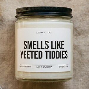 Puede incluir: Una vela en tarro de cristal con tapa negra y etiqueta blanca que dice "SMELLS LIKE YEETED TIDDIES". La vela está rodeada de un cortamechas negro, una caja de cerillas y flores secas. La vela es de 255 gramos.