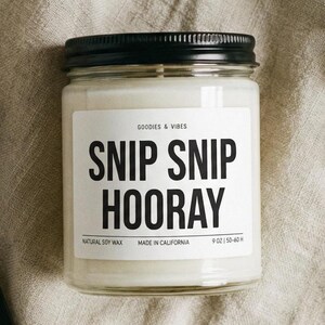 Puede incluir: Vela de cera de soja blanca en un tarro de cristal con tapa negra. La etiqueta dice "SNIP SNIP HOORAY". Un cortamechas negro y cerillas están junto a la vela. La vela es de 255 gramos.