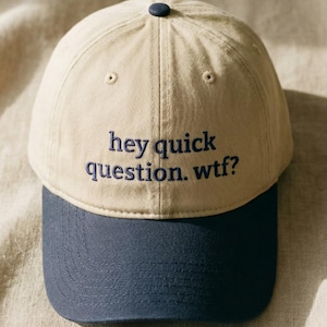 May include: A beige baseball cap with a navy blue peak. The cap has the embroidered text "hey quick question. wtf?" in navy blue. A small navy blue button is on top.