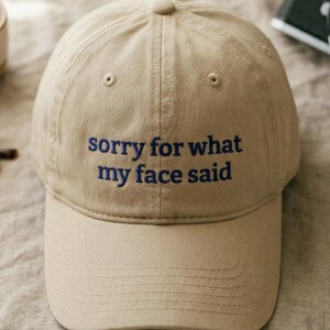 May include: A beige baseball cap with the embroidered phrase "sorry for what my face said" in blue. The cap features a curved peak and a button on the crown. A cup, sunglasses, and a camera are in the background.