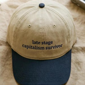 Puede incluir: Gorra de béisbol beige con visera azul marino. La gorra tiene el texto bordado "late stage capitalism survivor" en azul marino. Tiene una visera curva y un botón en la parte superior.