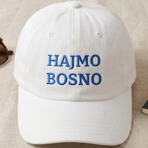 May include: A white baseball cap with the blue embroidered text "HAJMO BOSNO". The cap has a curved brim and a button on top. Sunglasses and a notebook are visible in the background.