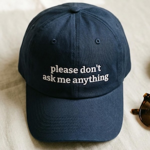 May include: A navy blue baseball cap with the text "please don't ask me anything" embroidered in white. The cap is on a cream-coloured surface, with sunglasses and a small plate nearby.