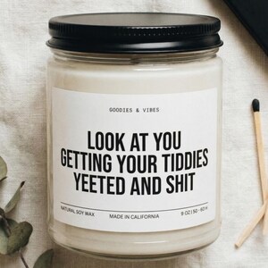 Puede incluir: Vela de cera de soja blanca en un tarro de vidrio con tapa negra. La etiqueta dice "LOOK AT YOU GETTING YOUR TIDDIES YEETED AND SHIT". Junto a la vela hay una caja de cerillas y una ramita de eucalipto.