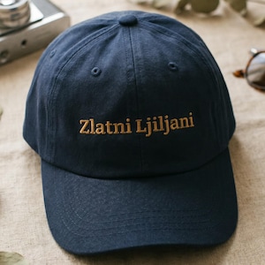 May include: A navy blue baseball cap with the embroidered text "Zlatni Ljiljani" in gold. The cap is made of a soft fabric and has a curved brim. The cap is a casual accessory.
