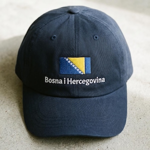 May include: A navy blue baseball cap featuring an embroidered flag of Bosnia and Herzegovina in yellow, blue, and white. The text "Bosna i Hercegovina" is embroidered in white thread directly below the flag on the front of the cap.