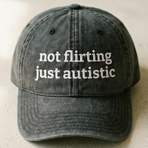 May include: A gray baseball cap with the embroidered text "not flirting just autistic" in white. The cap has a curved brim and a classic six-panel design, suitable for casual wear.