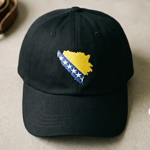 May include: A black baseball cap featuring an embroidered patch of the map of Bosnia and Herzegovina. The map is yellow with a blue diagonal stripe containing white stars. The cap has a structured crown, a curved brim, and a button on top.