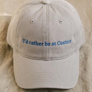 Puede incluir: Una gorra de béisbol de color beige claro con el texto azul bordado I'd rather be at Costco centrado en el panel frontal. La gorra tiene una corona estructurada, una visera curva y pequeños ojales de ventilación en los paneles superiores.