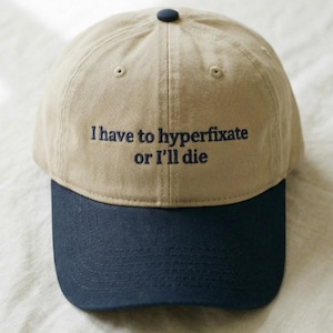 May include: A beige baseball cap with a navy blue peak. The cap has the embroidered text, "I have to hyperfixate or I'll die" in navy blue thread. The cap has a navy blue button on top.