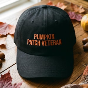 May include: A black baseball cap featuring the embroidered orange text PUMPKIN PATCH VETERAN on the front panel. The cap has a structured crown, a curved brim, and a button at the top center, constructed from a solid black fabric material.