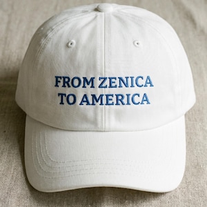 May include: A white baseball cap with the embroidered text "FROM ZENICA TO AMERICA" in blue. The cap has a curved brim and a button on top. The background is a neutral, textured fabric.