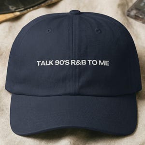 Könnte beinhalten: Marineblaue Baseballkappe mit dem weißen Text "TALK 90'S R&B TO ME" auf der Vorderseite gestickt. Die Kappe ist aus weichem Stoff und hat einen gebogenen Schirm. Im Hintergrund befinden sich eine Schallplatte und eine Kassette.