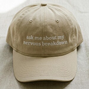 Könnte beinhalten: Beige Baseball Cap mit gebogenem Schirm. Der Hut hat den gestickten Text "ask me about my nervous breakdown" in Weiß. Der Hut ist aus einem weichen Stoff gefertigt und hat ein klassisches Sechs-Panel-Design.
