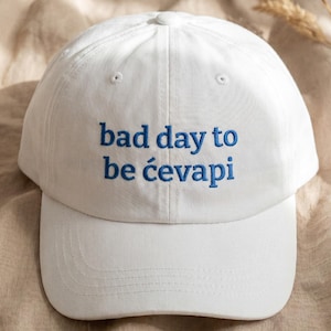 May include: White baseball cap with the blue embroidered text "bad day to be ćevapi". The cap has a curved brim and a fabric strap closure. The hat is made of a soft, durable material.