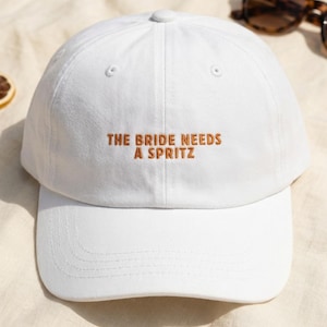 May include: A white baseball cap with the embroidered phrase "THE BRIDE NEEDS A SPRITZ" in brown. The cap is set against a light beige backdrop, with sunglasses and dried orange slices visible.