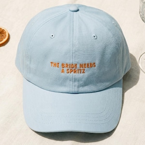 May include: A light blue baseball cap with the embroidered text "THE BRIDE NEEDS A SPRITZ" in orange. The cap has a curved brim and a rounded top. The background includes a glass and dried orange slices.