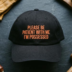 May include: A black baseball cap featuring embroidered orange text on the front that reads, PLEASE BE PATIENT WITH ME I'M POSSESSED. The cap has a structured crown, a curved brim, and eyelets for ventilation, constructed from a durable fabric.