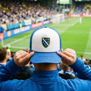 Könnte beinhalten: Eine weiß-blaue Trucker-Cap mit einem mittigen Wappenemblem, das einen diagonalen Streifen und gelbe Lilien zeigt. Die Kappe hat ein festes Vorderteil, ein Netzgewebe an der Rückseite und einen blauen Schirm vor einem Stadionhintergrund.
