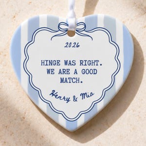 May include: A heart-shaped ceramic ornament featuring light blue and white vertical stripes. The center displays a white scalloped label with the text 2026, HINGE WAS RIGHT. WE ARE A GOOD MATCH. Henry & Mia, topped with a blue bow graphic.