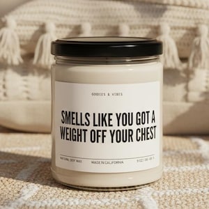 Op de afbeelding: Een glazen potkaars met een zwart deksel en een wit etiket met de tekst "Smells Like You Got A Weight Off Your Chest." De kaars is omgeven door decoratieve kussens en een kleine vetplant in een witte pot. De kaars is 255 gram.