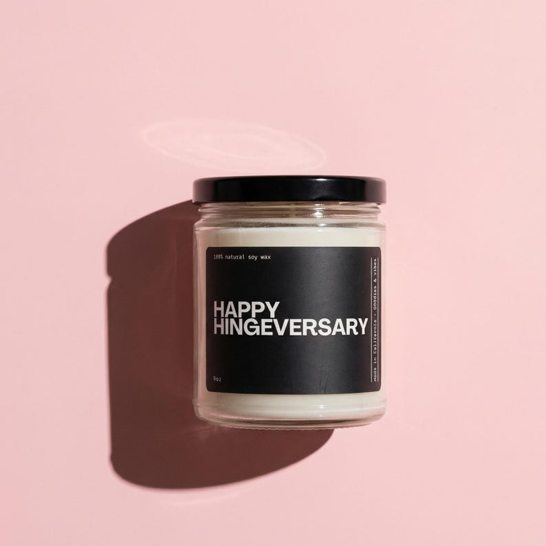 May include: A clear glass jar candle with a black lid and a black label that reads "HAPPY HINGEVERSARY." The candle is filled with white wax and sits on a pink surface. The label also states "100% natural soy wax."