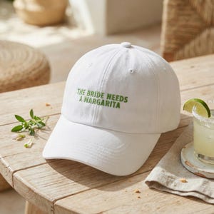 May include: White baseball cap with the text "THE BRIDE NEEDS A MARGARITA" embroidered in green. The cap is on a wooden surface, next to a margarita cocktail.