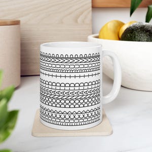 May include: White ceramic mug with a black geometric pattern. The design features various shapes and lines arranged in horizontal bands around the mug. The mug is sitting on a beige coaster.