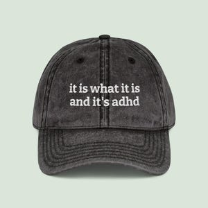 May include: A faded black baseball cap with a curved peak. The cap has the embroidered text "it is what it is and it's adhd" in white. The cap has a classic six-panel design and is a dark grey colour.