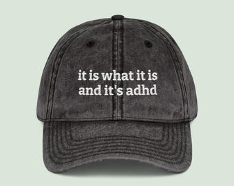 May include: A distressed black baseball cap with a curved brim. The cap features the embroidered text "it is what it is and it's adhd" in white, centered on the front. The cap has a classic six-panel design.