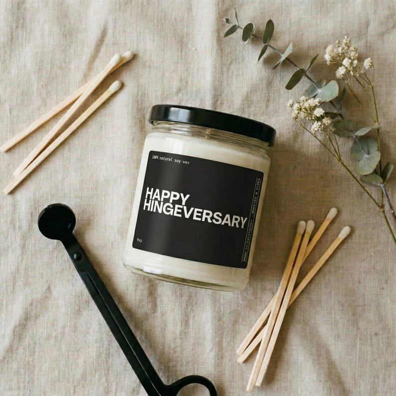 May include: A soy wax candle in a glass jar with a black label that reads "HAPPY HINGEVERSARY." The candle is surrounded by wooden matches, a black wick trimmer, and sprigs of greenery. The candle is 4 oz.