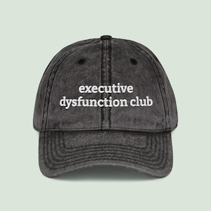 May include: A faded black baseball cap with a curved peak. The cap displays the embroidered text "executive dysfunction club" in white, positioned centrally. The hat has a traditional six-panel construction, a button on top, and an adjustable strap.