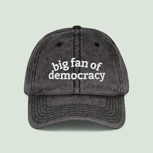 May include: A charcoal gray baseball cap with a curved brim. The cap features the embroidered text "big fan of democracy" in white, centered on the front. The hat has a vintage, worn look.