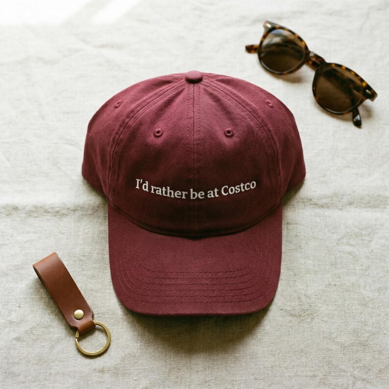 Op de afbeelding: Bordeauxrode baseballcap met de geborduurde tekst "I'd rather be at Costco". De pet ligt naast een zonnebril met schildpadmotief en een bruine leren sleutelhanger met een goudkleurige ring.