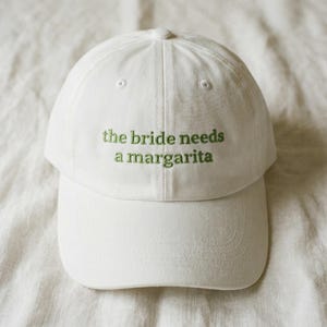 Op de afbeelding: Witte baseballcap met de groen geborduurde tekst "the bride needs a margarita". De cap heeft een gebogen klep en een knoop aan de bovenkant. De hoed is gemaakt van een zachte stof.