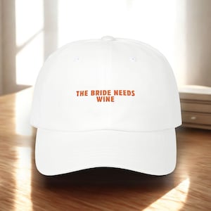 May include: White baseball cap with the text "THE BRIDE NEEDS WINE" in orange letters. The cap has a curved brim and a button on top. The hat is sitting on a wooden surface.